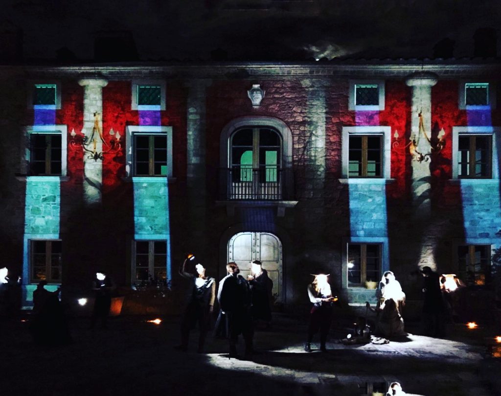 Scena di "Rodrigo", spettacolo per la regia di Gabriele Valentini. In foto attori sparsi per la scena, alle loro spalle una casa su cui sono proiettate immagini di colonne.