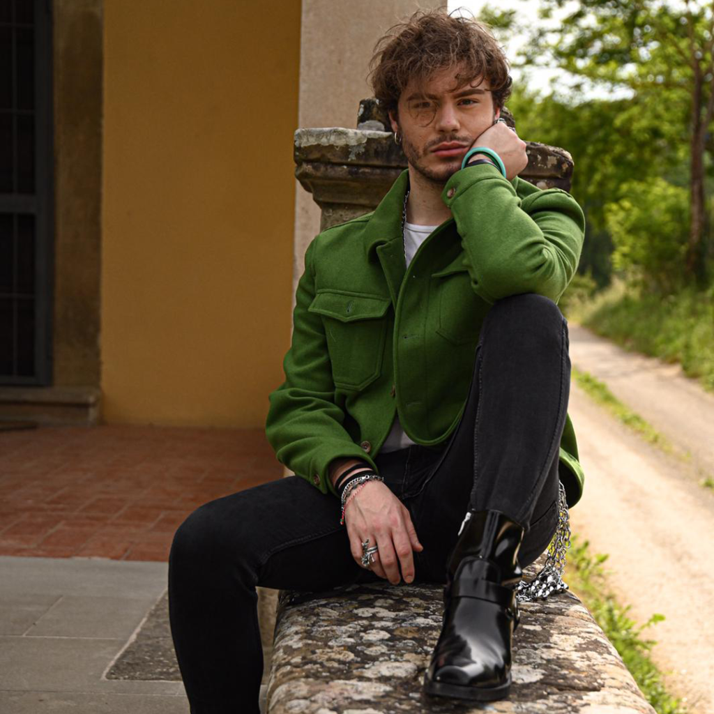 Luca Gremita, seduto su un muretto, indossa una giacca verde e dei pantaloni neri.