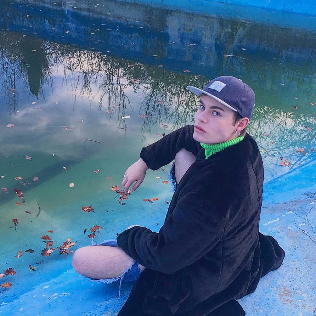 Luca Gremita, seduto sul bordo di uno specchio d'acqua, indossa un lungo cappotto nero. L'immagine è tutta sui toni dell'azzurro.