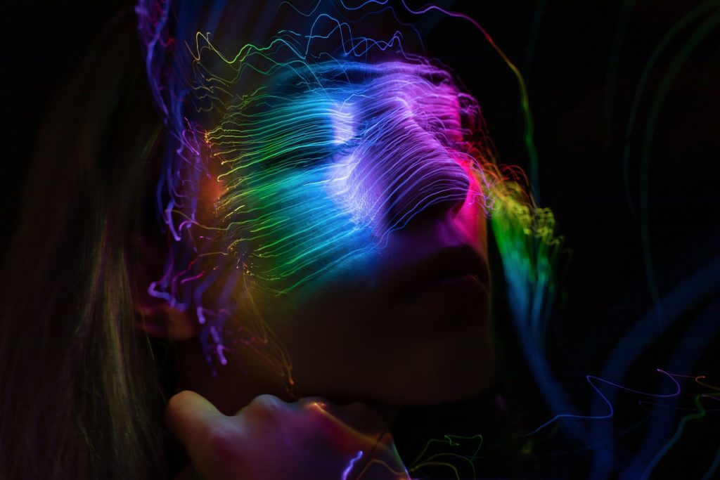 Primo piano di una persona con capelli lunghi biondi in un ambiente buio: sulla sua faccia passano delle luci arcobaleno.
